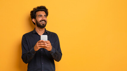 Dreamy Young Indian Guy Holding Smartphone And Looking Aside At Copy Space