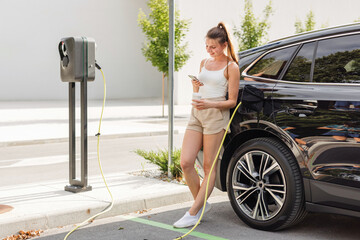 Young woman charging her elegant black EV car at a public electric station in the neighborhood....