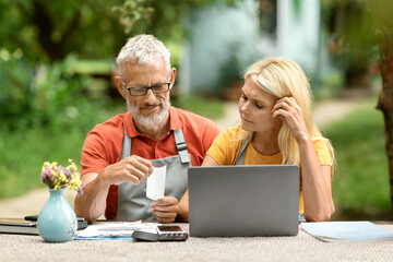 Obraz premium Stressed senior couple with laptop checking bills, sitting at table in garden