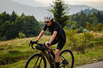 Fototapeta premium Woman cyclist wearing black cycling kit. She is riding a gravel bike on a gravel road at sunset with a view of the mountains.Empty mountain road.Cycling gravel adventure in Romania.Bucegi Natural Park