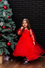 Little girl in red dress smiling by Christmas tree. Little beautiful girl in a red evening dress the Christmas tree. Girl in red in Christmas decorations. Christmas concept. New Year. Baby near a