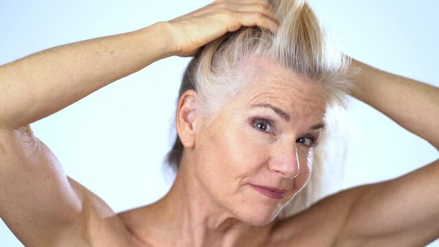 Beautiful middle-aged woman examines her blond hair turning grey in the mirror. She cares about gray hair.