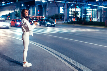 Young woman standing on the street in the night holding smartphone, waiting for taxi, full length, copy space