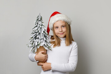 Young adorable child girl in Santa Claus hat posing with Christmas tree in her hands on white background, Christmas portrait