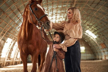 Woman with her little daughter is with horse indoors
