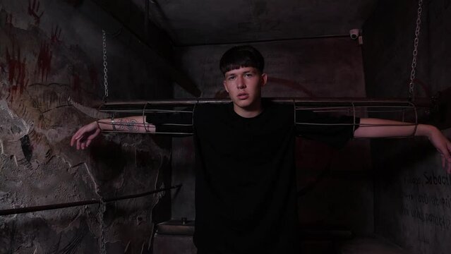 A young man is chained to horizontal handcuffs in a dark quest room. A dark prison room with old rusty walls and antique shackles. The room of horrors.