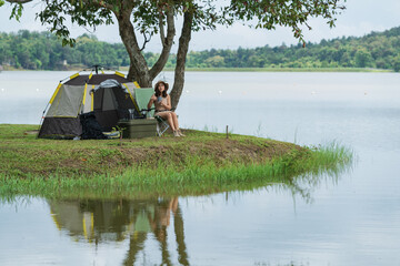 An Asian woman is enjoying sitting near a lake and drinking hot coffee in nature while in a tent camp outdoors camping.