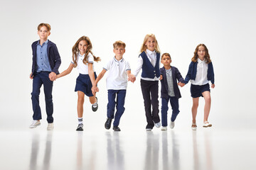 Obraz premium Group of stylish children, boys and girls, pupils in school uniform running against white studio background. Classmates. Concept of childhood, school, education, fashion, style. Copy space for ad