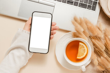 Woman hold smartphone with white screen closeup, laptop background with tea, candle, lagurus grass on linen cloth, aesthetic home workplace. Website promotion, business branding, autumn theme