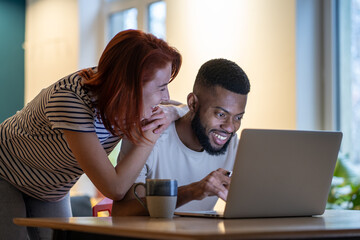 Loving young caucasian woman talking with boyfriend working on laptop late at home, smiling girlfriend demanding attention from busy with work African American guy freelancer. Work life balance
