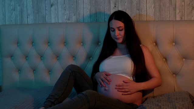A pregnant woman sits on the bed, gently caressing