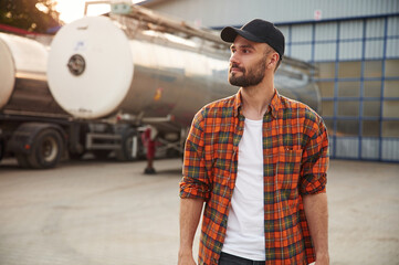 Sunny day, standing against fuel tanker. Young truck driver in casual clothes