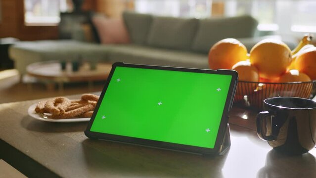 Tablet computer with green screen chromakey display with motion trackers lay on table near oranges, cookies and cup of hot tea and sofa on background.cozy interior of living room in house on daylight