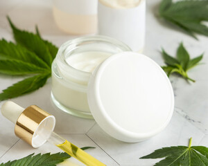 Obraz na płótnie Canvas Cream jar with a blank lid and pipette with CBD oil near cannabis leaves. Mockup