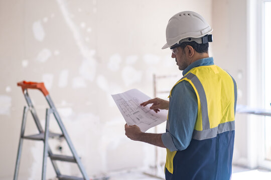 Professional engineer in white hardhat looking at project drawing in hands standing in room against ladder near unfinished wall renovation process in apartment