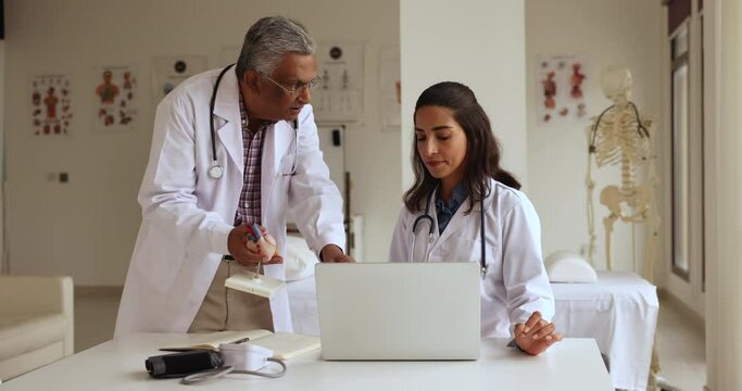 Indian and Latina cardiologist colleagues work together in clinic office, using laptop discuss telemedicine platform, old experienced GP teach intern at meeting in hospital. Internship, apprenticeship