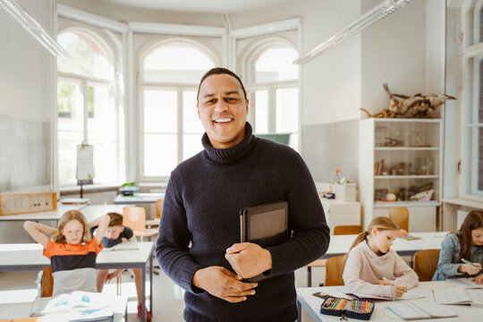 Portrait of smiling male teacher holding tablet PC while standing in classroom