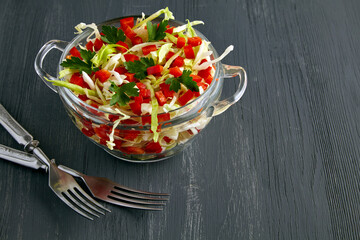 Light vegetable salad of kale and bell peppers in a glass bowl and cutlery on a black wooden table