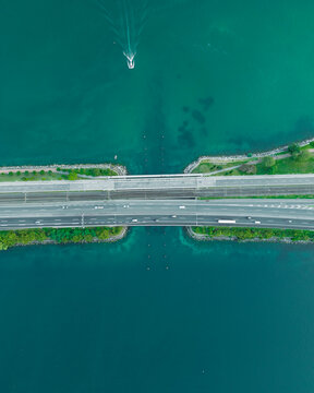 Aerial view of vehicles driving on a road crossing the Lugano lake with a bridge and a boat sailing under the bridge, Melide, Ticino, Switzerland.