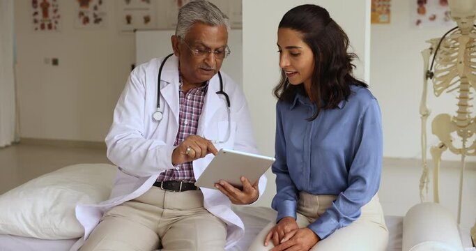 Indian mature GP explain treatment plan, showing check up health test results on digital tablet to patient at visit in clinic, client receive professional medical services, talk to doctor in uniform