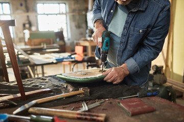 Closeup of senior man repairing old furniture in carpentry workshop and holding electric drilling...