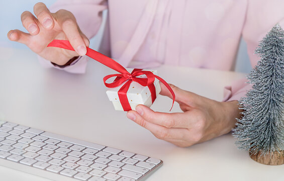 First person top view photo of young woman's hands unpacking small white gift box with red dotted ribbon bow on office white background