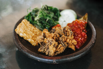 A wooden plate of Indonesian traditional food called Lalapan contains Belut Goreng or fried eel, vegetables, fried tempe, cucumber and egg plant with sambal or chili. Selective focus.