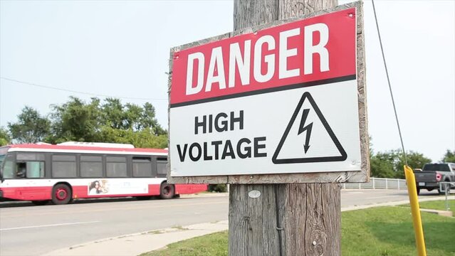 danger high voltage sign on wood post with bridge road behind with bus vehicles traffic cars passing on it, red white black with bent arrow illustration in triangle