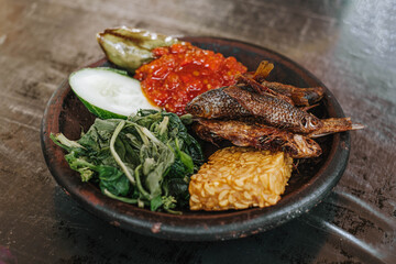 A wooden plate of Indonesian traditional food called Lalapan contains fried Iwak Wader or Spotted Barb fish, vegetables, fried tempe, cucumber and egg plant with sambal or chili. Selective focus.