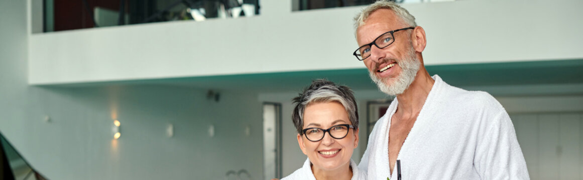 Joyful Middle Aged Couple In White Robes And Glasses Looking At Camera In Spa Center, Banner