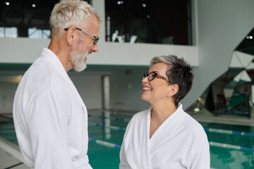 joyful middle aged couple in glasses and white robes chatting near indoor pool in spa center