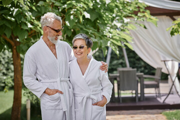 happy mature couple in sunglasses and robes standing in luxury resort, wellness retreat concept