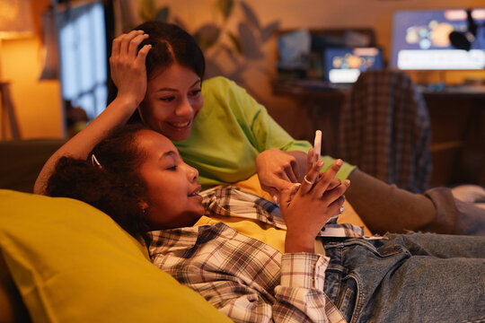 Side view portrait of happy black mother with teen daughter lying on bed together and using smartphone in cozy warm light