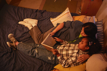 Portrait of young African American mother and daughter lying in bed together and reading book at night