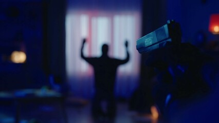The arrest of a burglar in a dark apartment lit by blue light. The offender is on his knees, with his hands up. A gun in the hand of a policeman on a blurred background of a criminal close up.