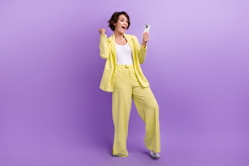 Full length photo of pretty lucky woman dressed yellow jacket winning game modern gadget isolated purple color background