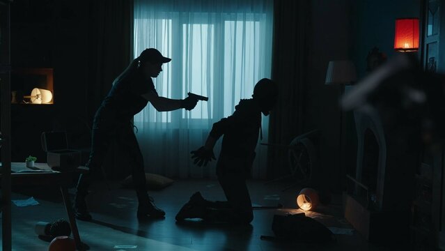 The arrest of a robber in a dark apartmen. Side view of a criminal in a black balaclava, kneeling at gunpoint with his hands behind his back. A female police officer holds the villain at gunpoint.