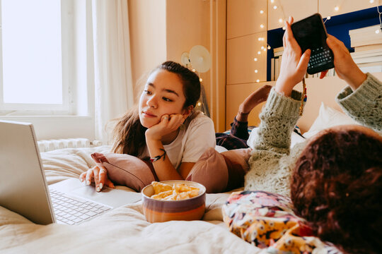 Young woman using laptop while friend text messaging through smart phone on bed
