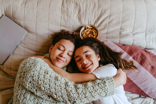 Smiling female friends embracing while lying on bed at home