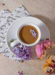 A cup of coffee with a pink donut on a light background with lilac flowers.