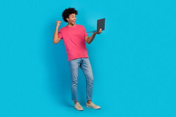 Full length photo of nice young guy hold device netbook raise fist winning wear trendy pink garment isolated on blue color background