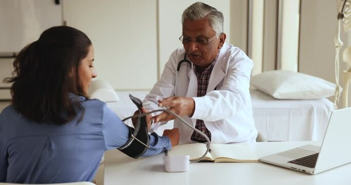 Mature Indian cardiologist apply cuff, use tonometer device to measuring blood pressure, pulse rate of female patient during visit at hospital, to determine state of cardiovascular system. Cardiology