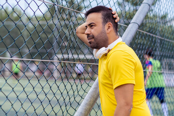 Worried man with wireless earphones watching football club training on large field leaning on metal...