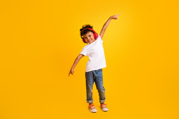 Full length photo of cheerful cool little boy dressed white t-shirt pointing fingers enjoying songs earphones isolated yellow color background