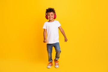 Full length photo of funky cheerful little boy dressed white t-shirt walking enjoying songs earphones isolated yellow color background