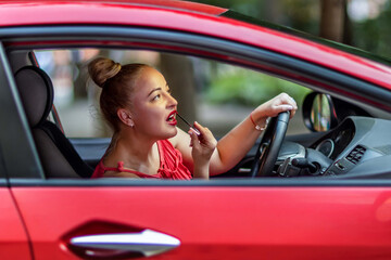 A woman in red paints her lips with lipstick, sitting at the wheel of a car, looking in the rearview mirror.