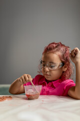 A beautiful little girl with crimson hair, wearing a pink shirt and safety glasses is playing educational games at the table