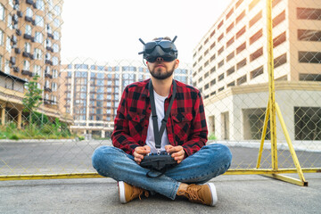 Portrait of a casually dressed young bearded man wearing goggles and using remote controller to operate fpv drone while sitting at the sreet road