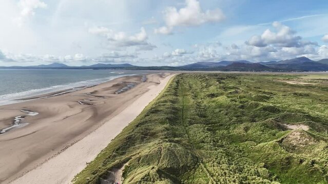 4K 60FPS aeriel shot of a sea and sandy beach shoreline in wales, uk, with sand dunes covered in long green gress on a hot summers day, mountains in the background