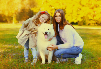 Happy family, young mother and child daughter with white Samoyed dog together in sunny autumn park